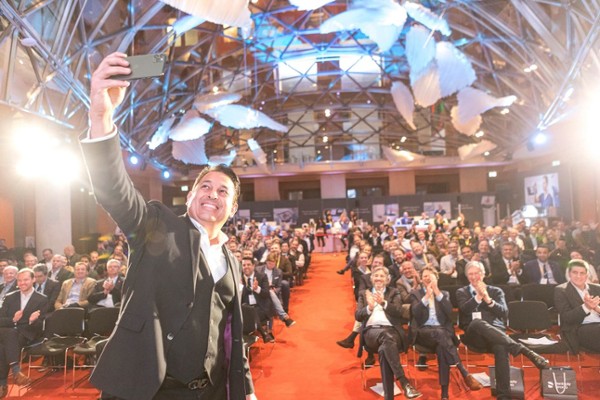 lachender Mann steht vor Saal voll mit Menschen, die auf Stühlen sitzen und klatschen, und macht ein Selfie