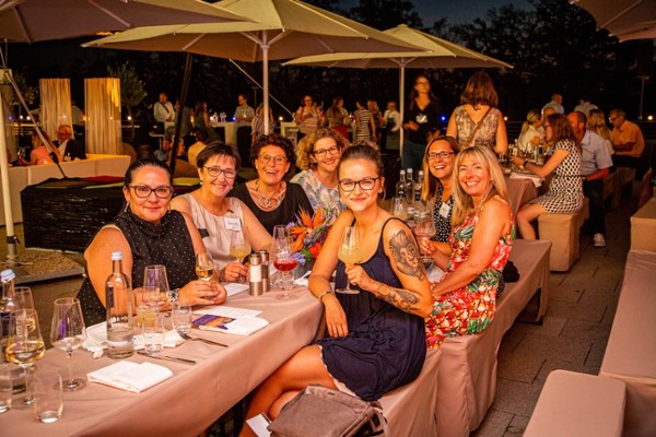 Eine Gruppe von Frauen sitzt draußen an einem langen, feierlich gedeckten Tisch und sind alle zur Kamera gedreht, im Hintergrund sind noch mehr Tische, an denen Menschen sitzen, zu sehen