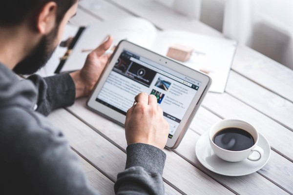 Blick über die Schulter eines Mannes, der an einem Tisch sitzt, auf den Bildschirm eines Tablets, rechts neben ihm steht eine volle Kaffeetasse, unscharf im Hintergrund liegen aufgeschlagene Zeitschriften