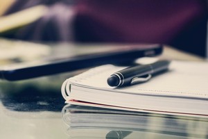 Frontalaufnahme eines Stifts auf einem Spiralblock liegend, dieser liegt auf einem Tisch, im Hintergrund ist unscharf ein Handy zu sehen